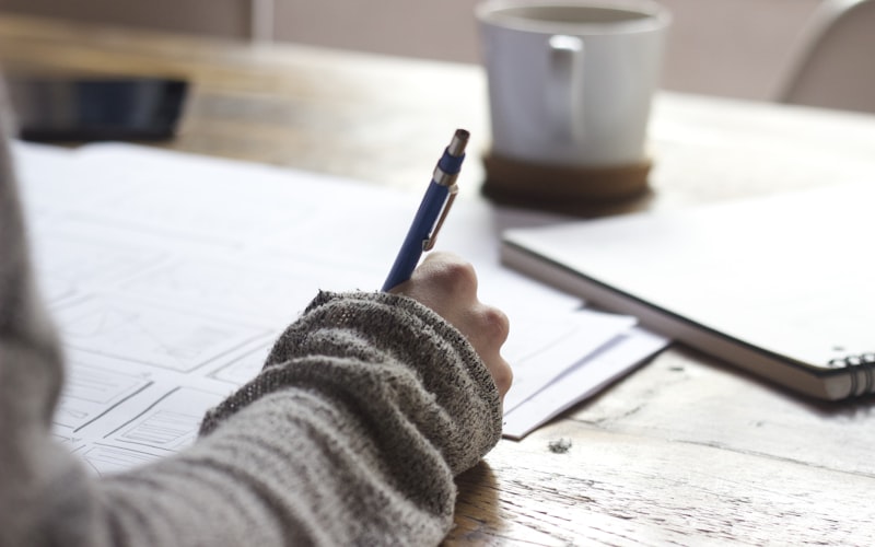 A hand writing notes at a warm timber desk beside a coffee cup